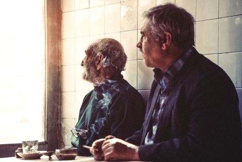 Free Two elderly men sitting in a Tehran café, one smoking, gazing out the window. Stock Photo