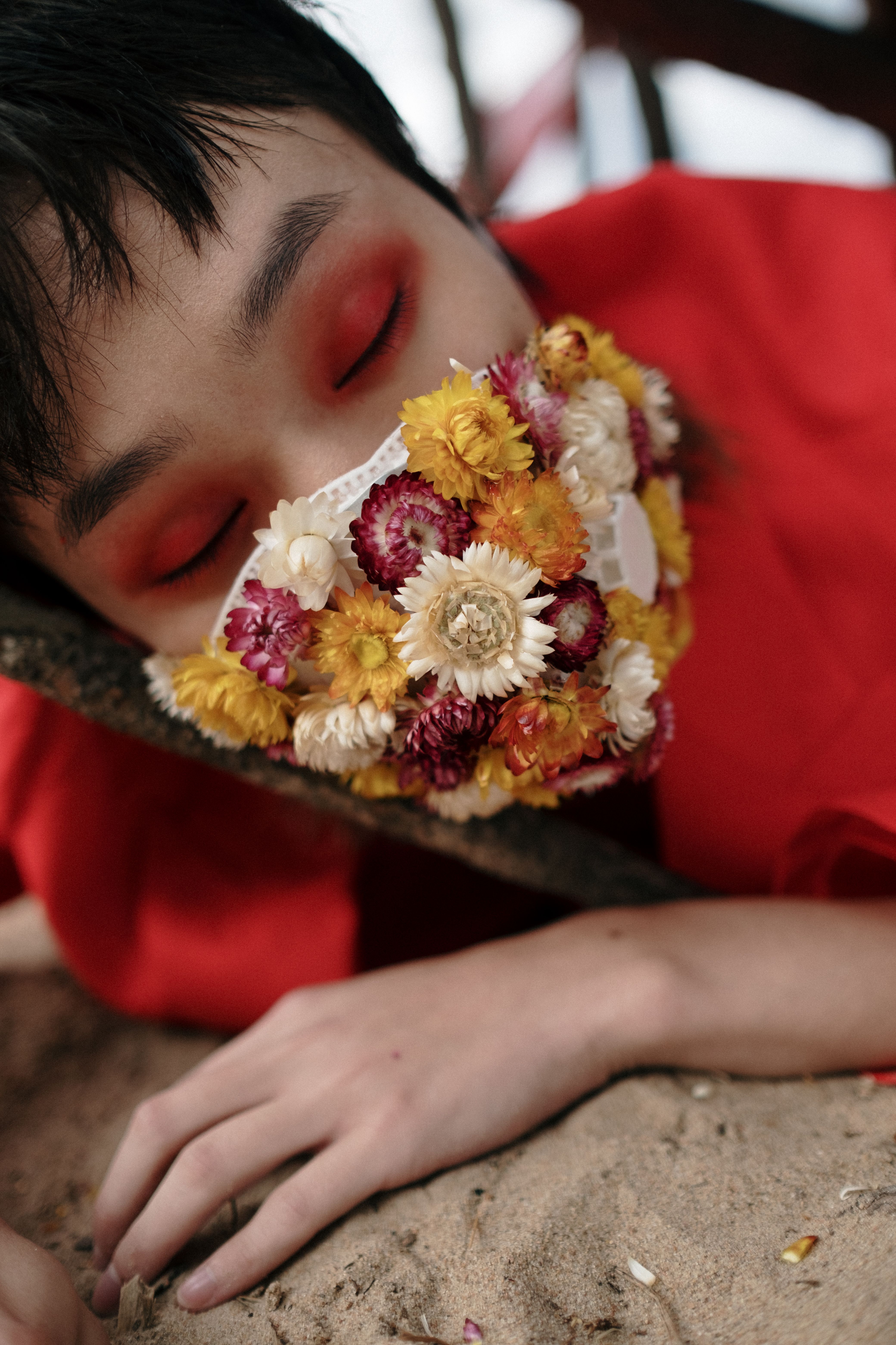 A creative photo of a person with eyes closed wearing a floral face mask on sand.