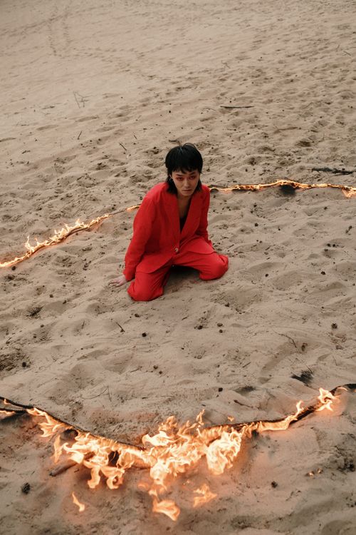 Free A person in a red suit kneels on a sandy beach, encircled by fire, creating a dramatic and conceptual scene. Stock Photo