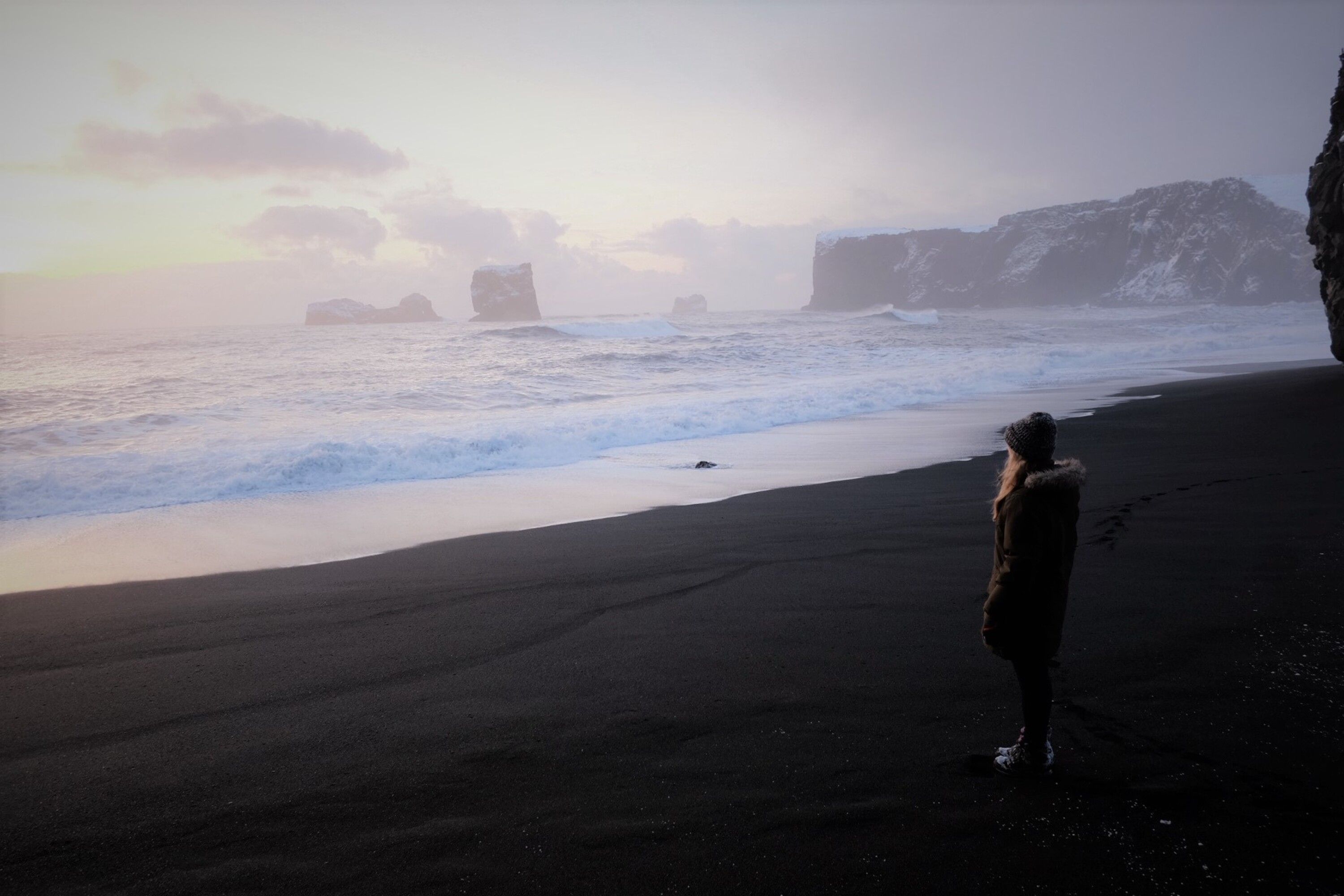 A woman in warm clothing stands on a black sand beach in Iceland, gazing at the ocean waves.