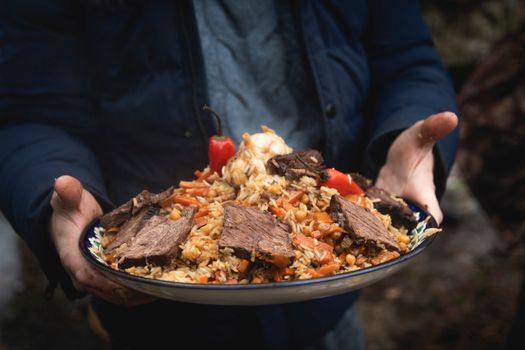 Close-up of a person holding a plate of beef pilaf with vegetables outdoors.