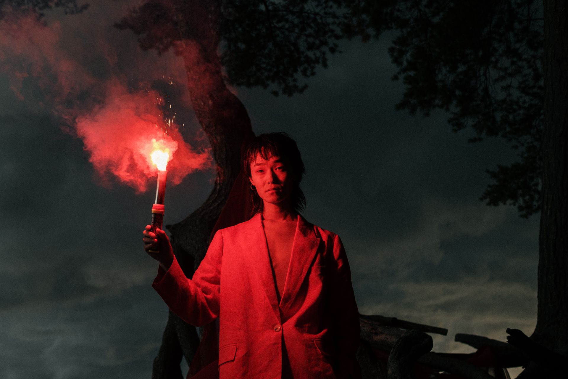 Asian man holding red flare in a forest at dusk, creating a dramatic moody atmosphere.