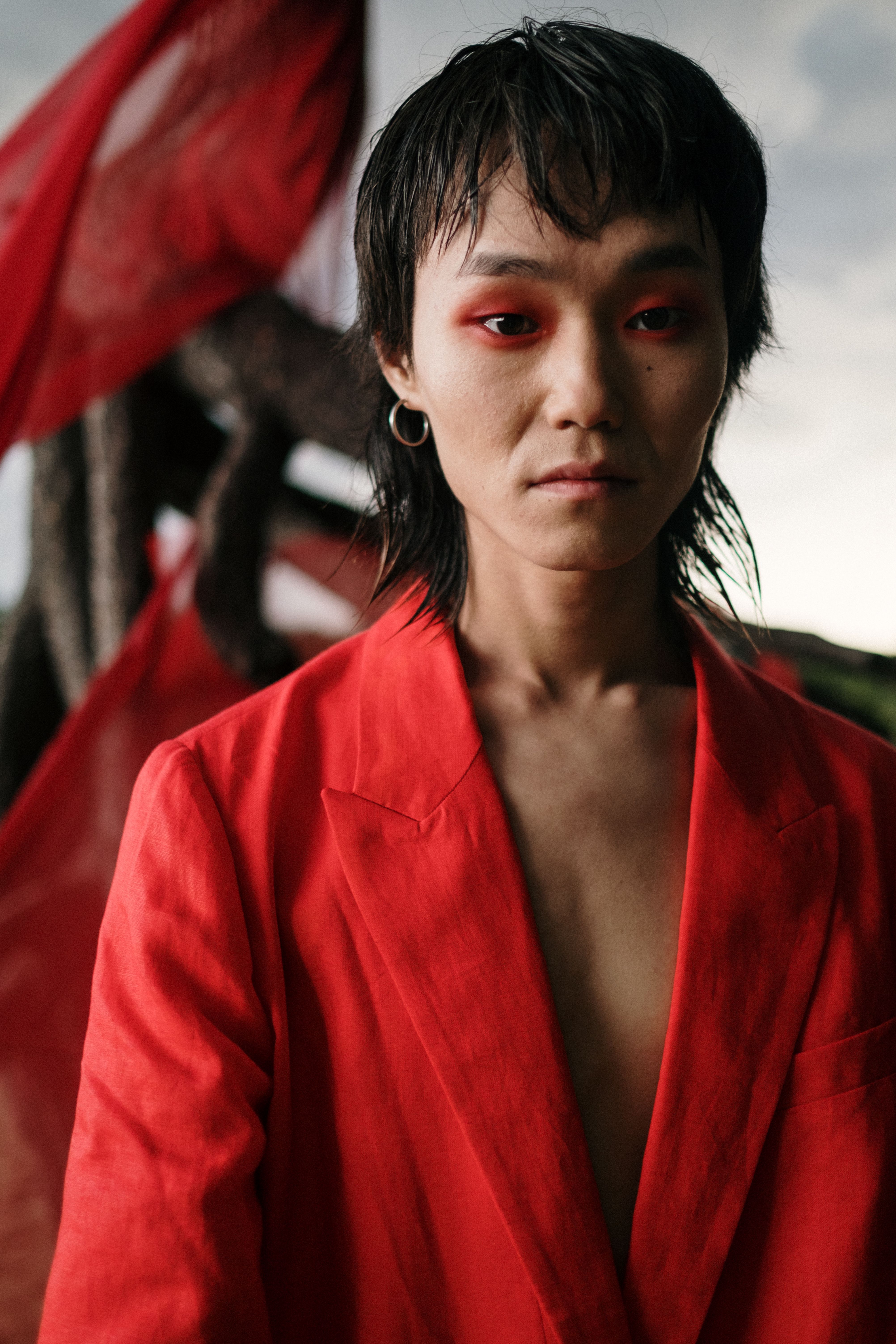 Androgynous model in red blazer with expressionless look, set against an outdoor background.