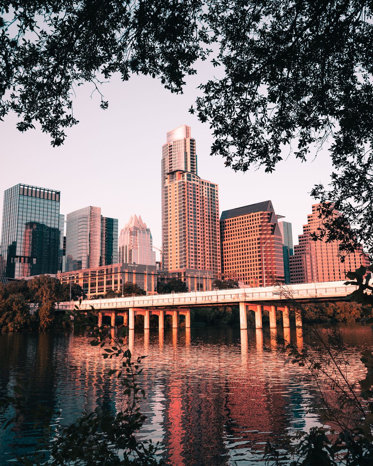 A beautiful view of the Austin skyline reflected in the river at sunset with a bridge and trees.
