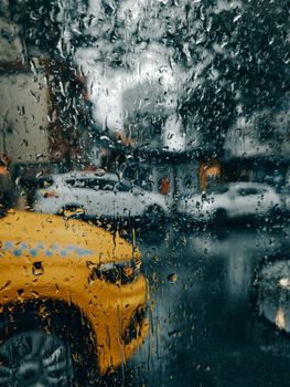 A rainy city street scene viewed through a window with raindrops, featuring cars and a yellow taxi.