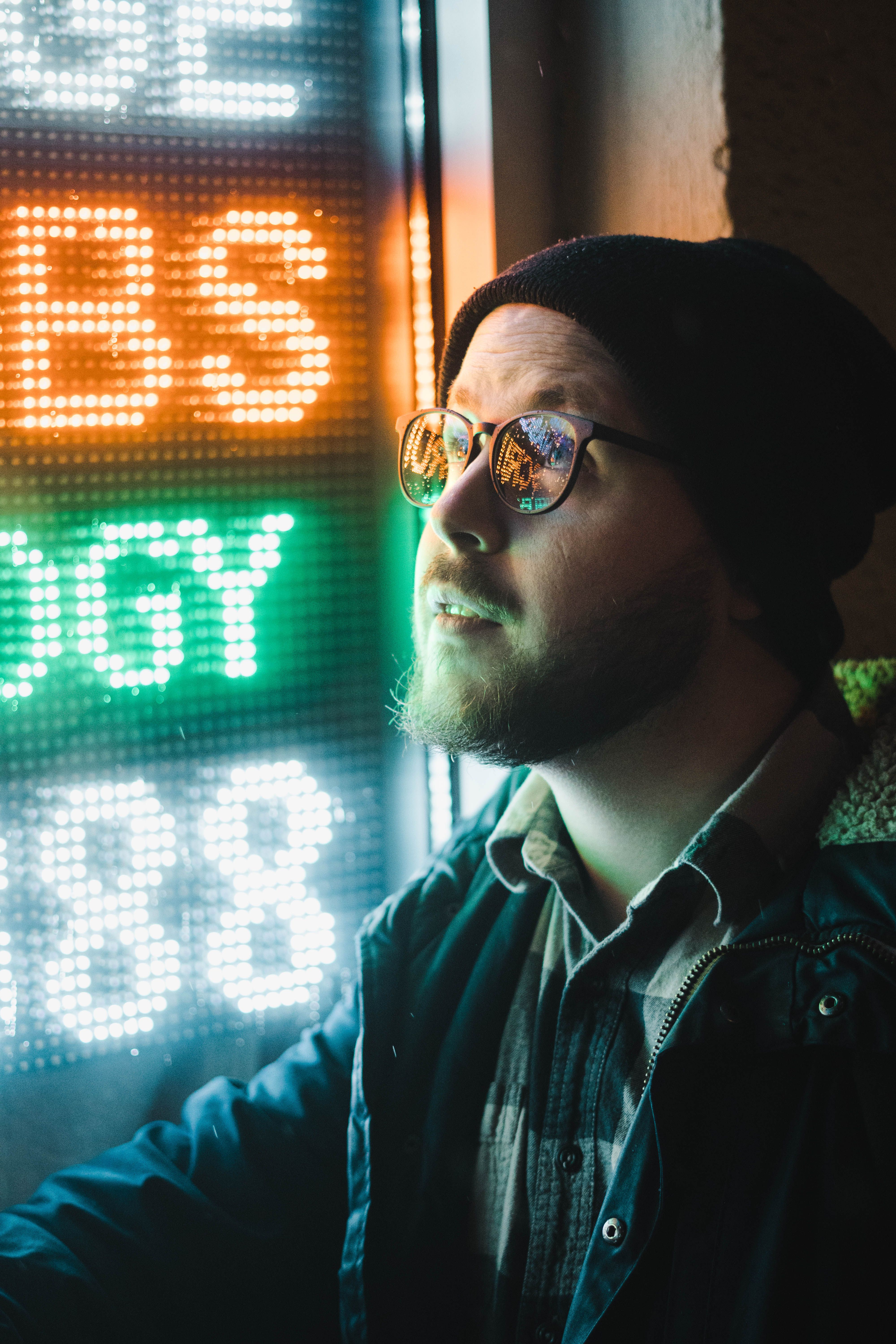 Artistic portrait of a man in a beanie gazing thoughtfully at a neon sign in Portland at night.