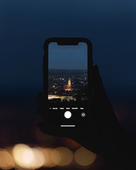 Silhouette of hand holding smartphone capturing city at night with glowing skyline.