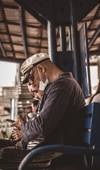 A man in a hat and face mask sits on a bench, focused on his smartphone.
