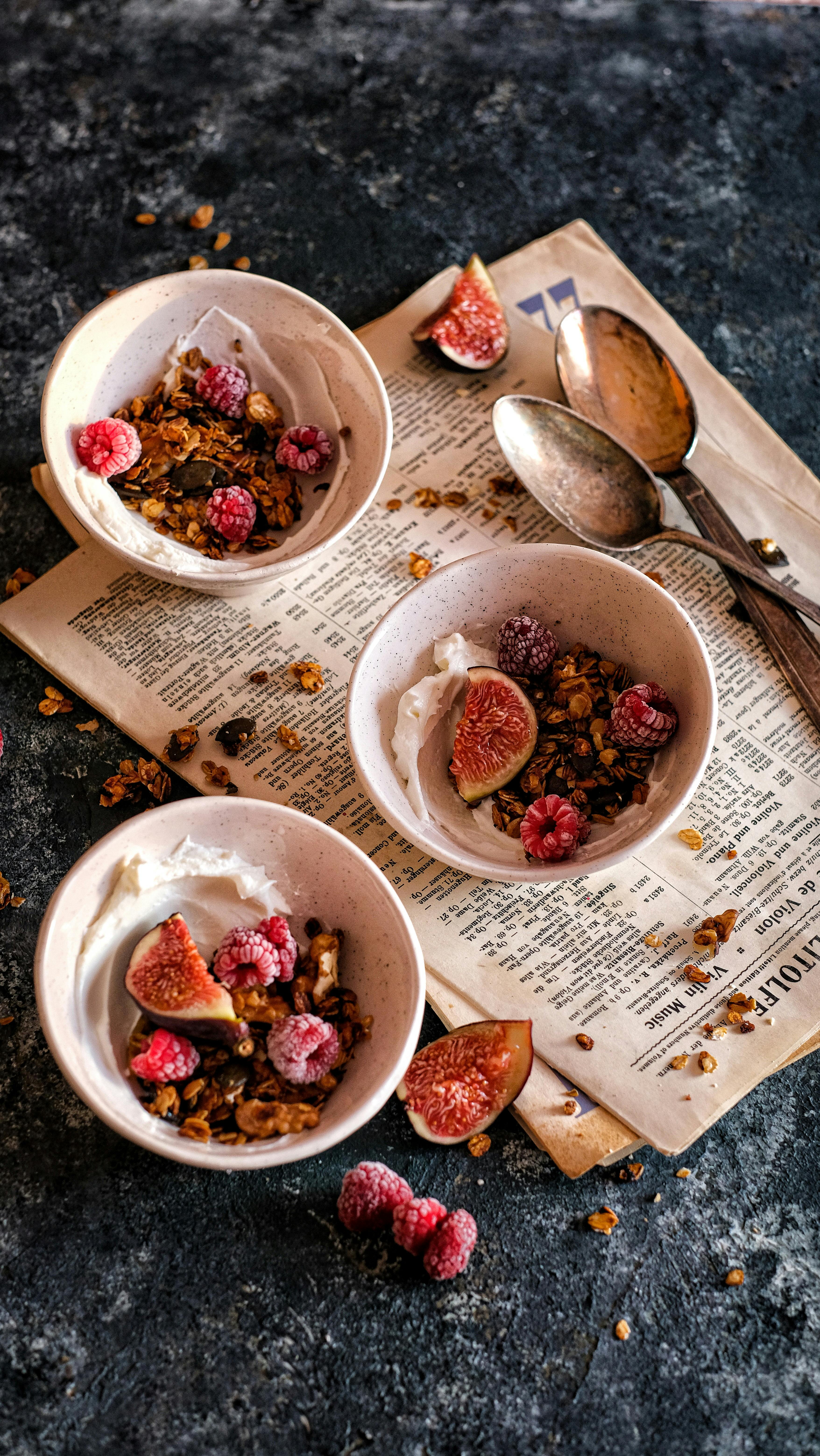 Three yogurt bowls with figs, raspberries, and granola on a rustic surface.