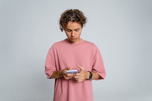 Teenage boy in pink shirt focuses on smartphone against gray background.