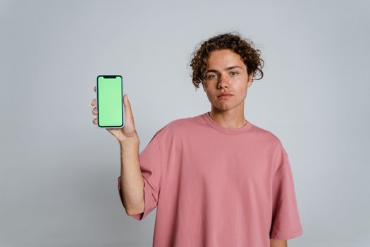 Portrait of a young man with curly hair holding a smartphone with a blank green screen in a studio setting.