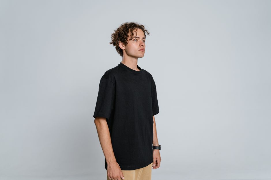 Side view of a teenager with curly hair wearing a black t-shirt and pants, standing against a white background.