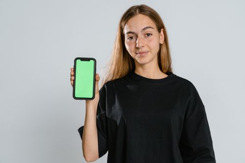 Free Young woman in black t-shirt showing a green screen smartphone in studio setting, ideal for mockup use. Stock Photo