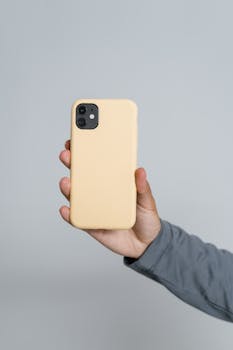 Close-up of a hand holding a smartphone with a yellow case on a neutral background.