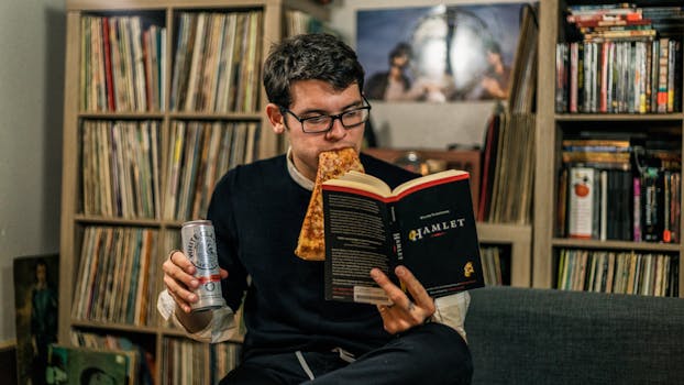 A man reads Hamlet while eating pizza and holding a drink in a cozy room.