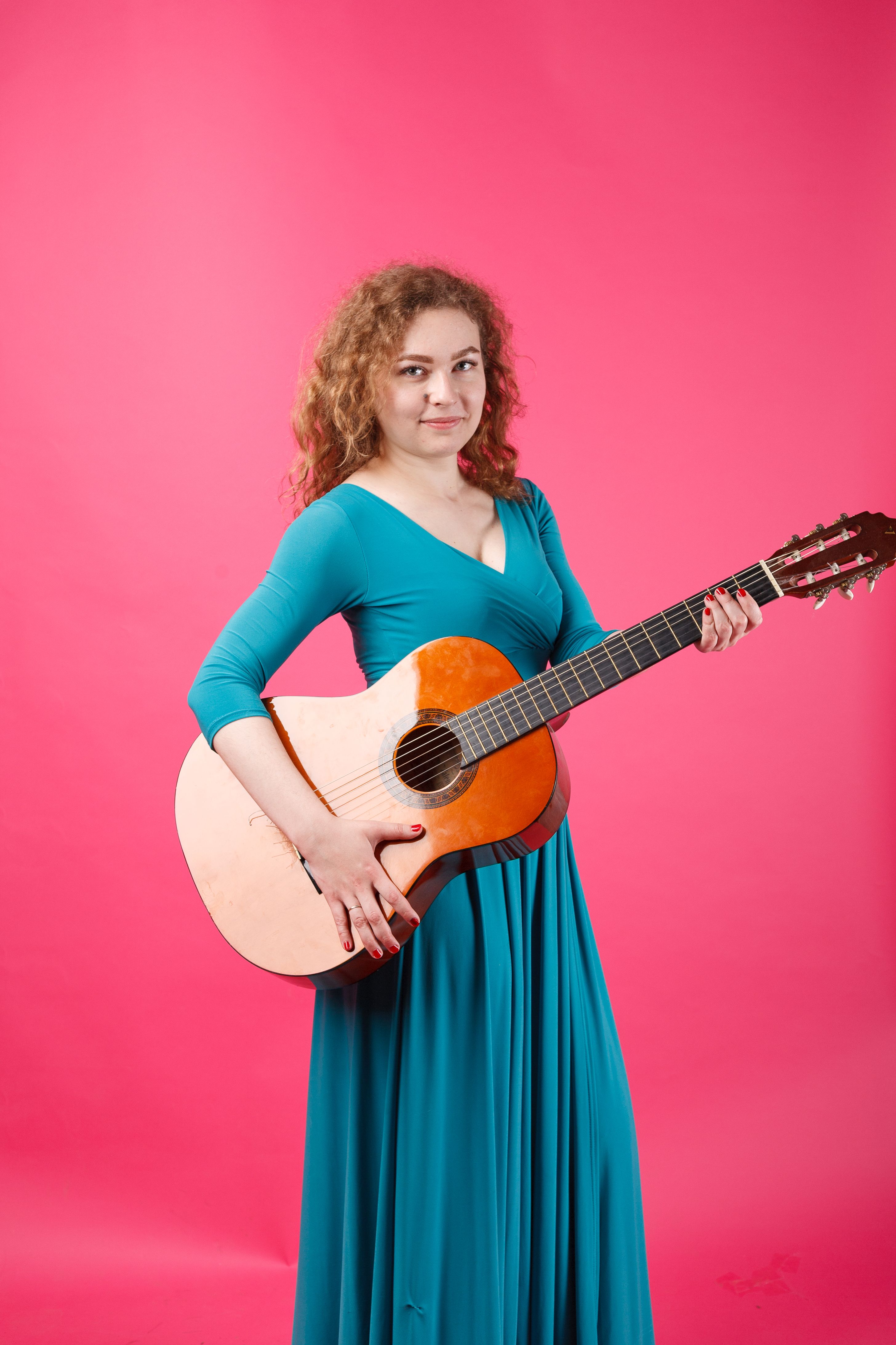 Woman in teal dress holding an acoustic guitar against a pink background.