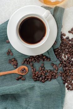 A top view of a cup of black coffee on a textured cloth with scattered roasted coffee beans.