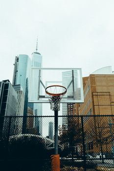 A basketball hoop in a city setting with skyscrapers in the background, framed by a chain-link fence.