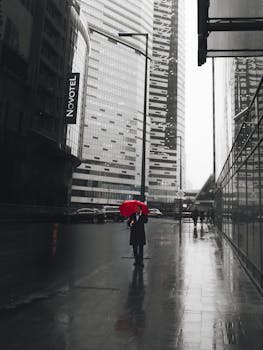 A striking urban photo featuring a lone person with a red umbrella amidst skyscrapers on a rainy day.