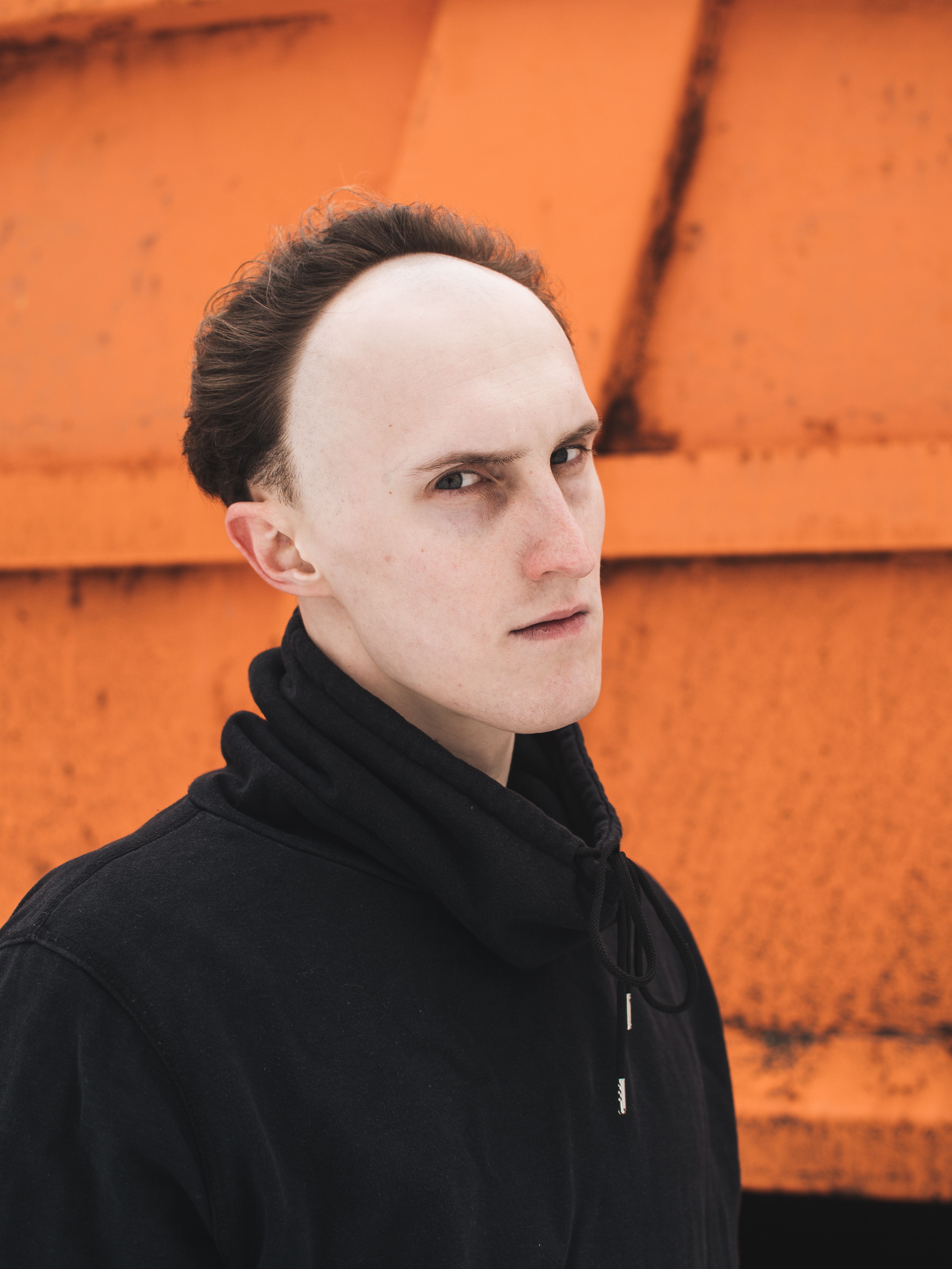 Close-up portrait of a man in black sweater with an orange industrial backdrop.