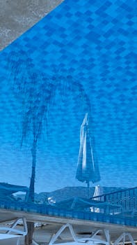 Captivating image of a patio umbrella reflecting in a pool, with a palm tree and mountain backdrop in Kaş, Turkey.