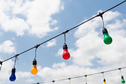 Vibrant string lights in blue, red, yellow, and green colors set against a bright sky with clouds.