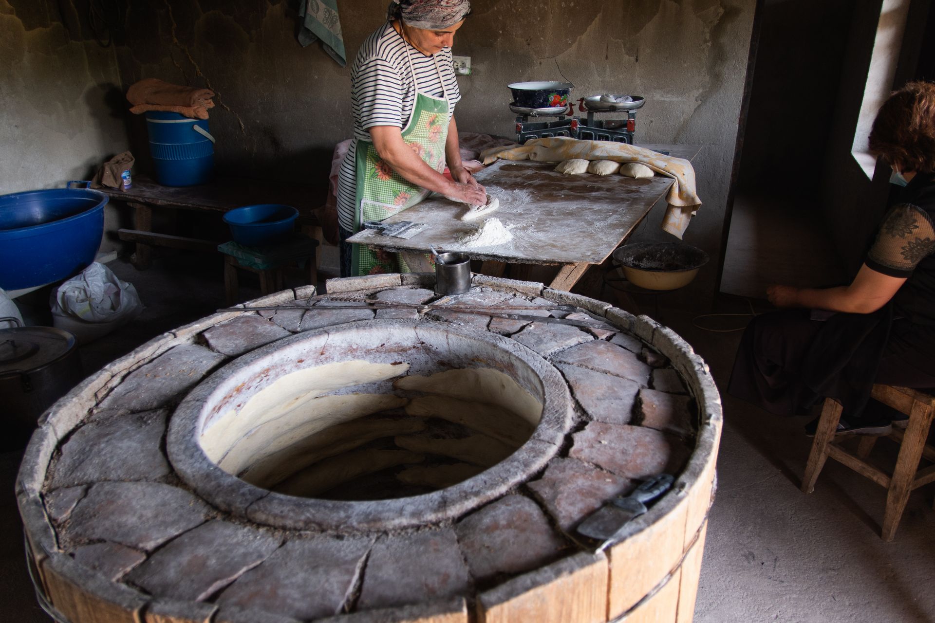 A woman preparing dough in a traditional kitchen with a clay oven nearby.