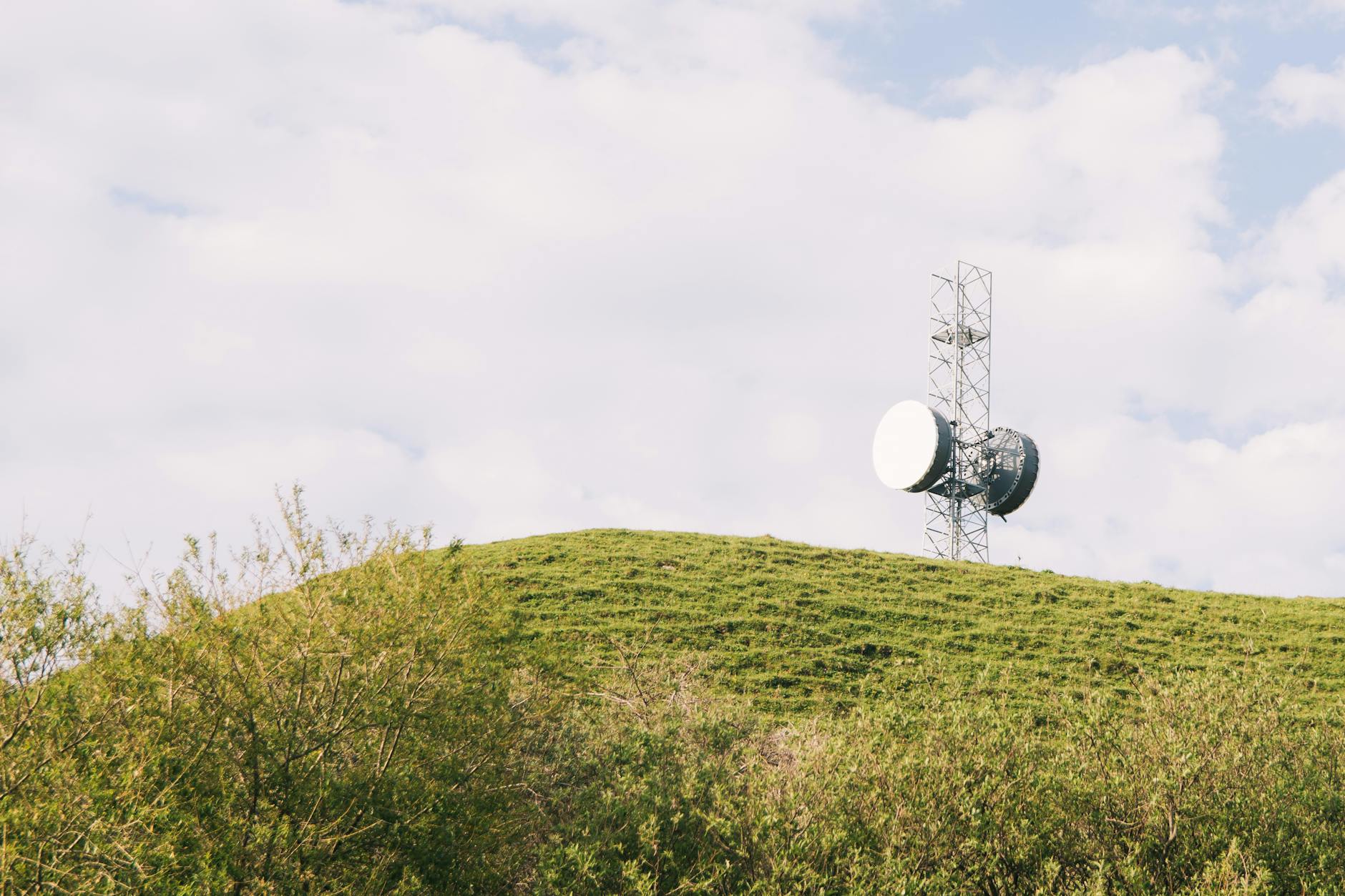 A communication tower on a grassy hill under a partly cloudy sky. Ideal for technology imagery.