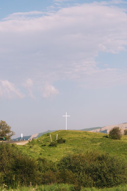 Free A tranquil rural scene featuring a white cross atop a grassy hill under a partly cloudy sky. Stock Photo