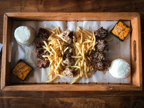 Gourmet meal of chicken wings, fries, rice, and sauces on a wooden tray, captured from above.