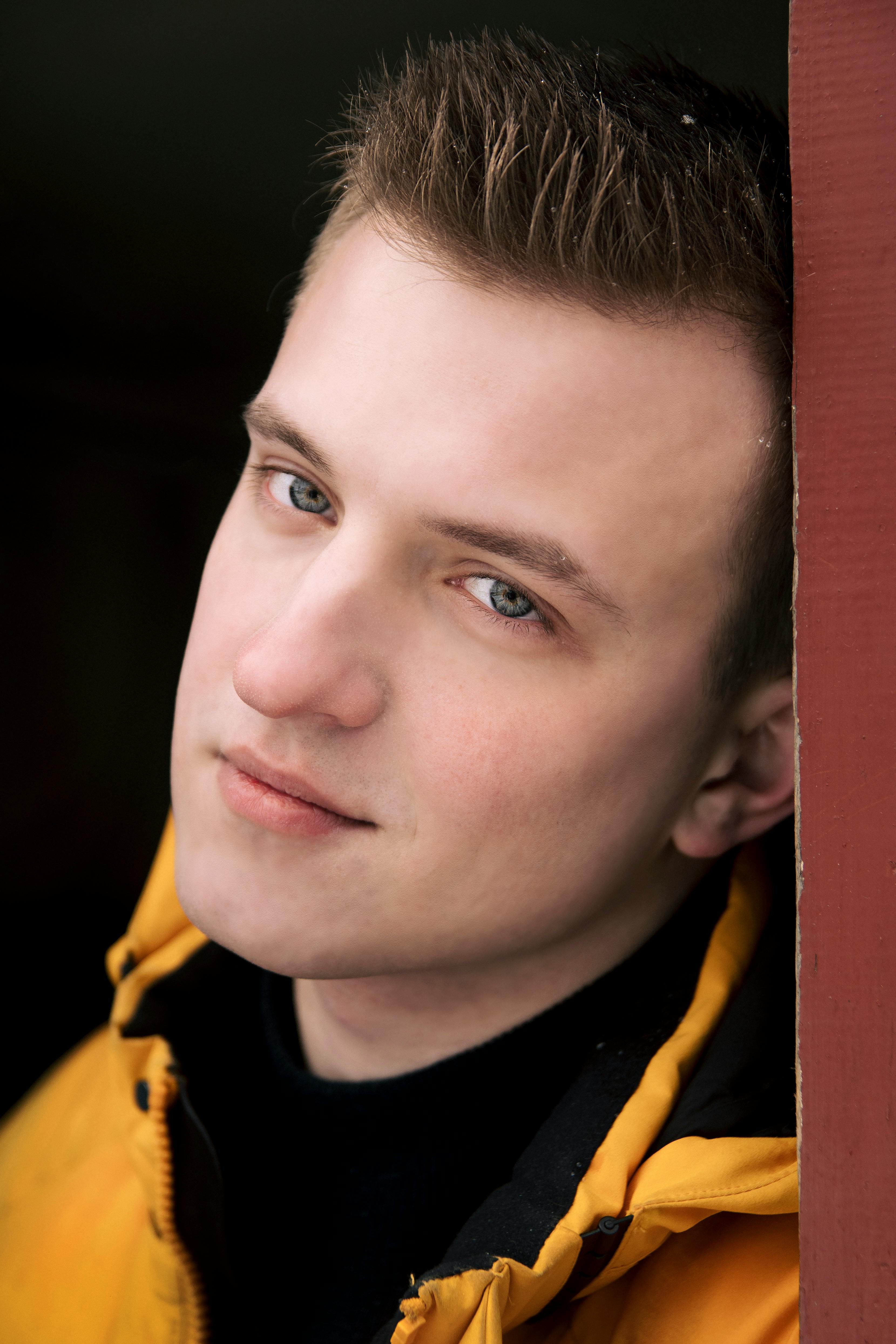 Close-up portrait of a young man in a yellow jacket, looking at the camera.
