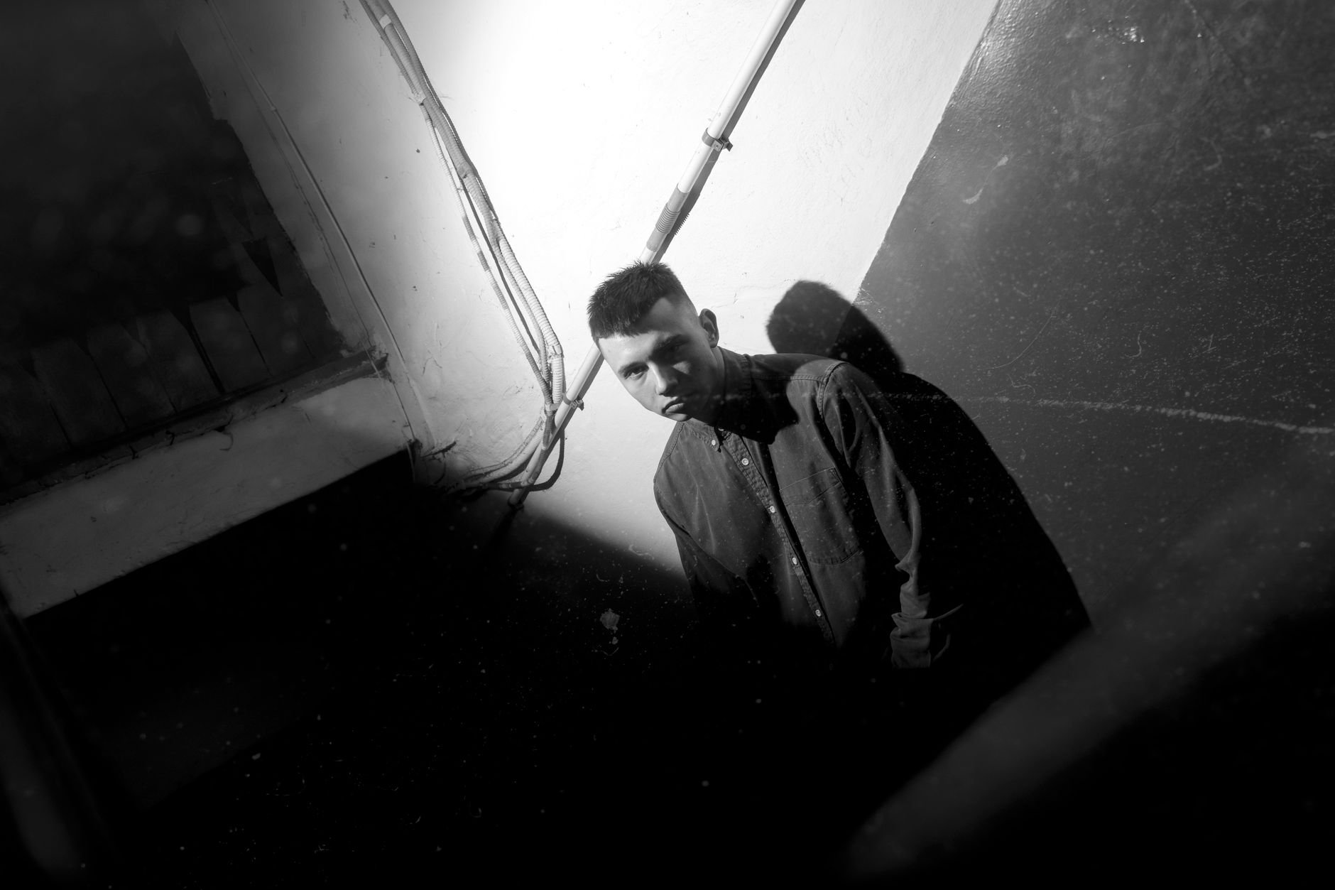 A serious man in shadow leaning against a wall in black and white, lit from one side