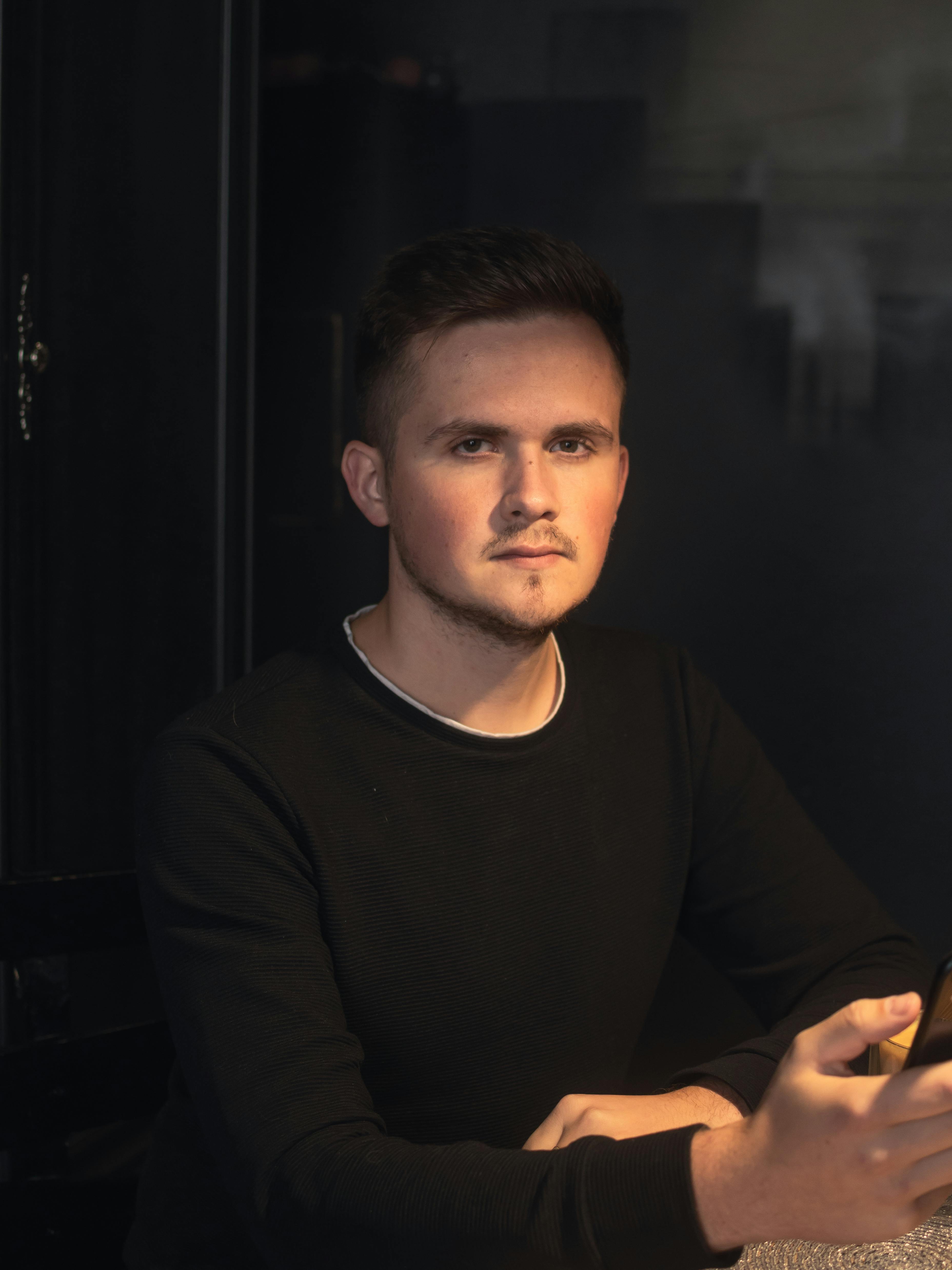 Portrait of a young man in dim light, wearing a black sweater, looking contemplative.