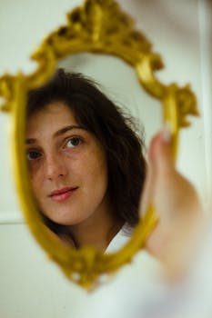 A woman gazes into an ornate gold mirror, reflecting her thoughtful expression.