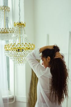 A woman with curly hair poses indoors with elegant chandeliers in the background, showcasing interior design.