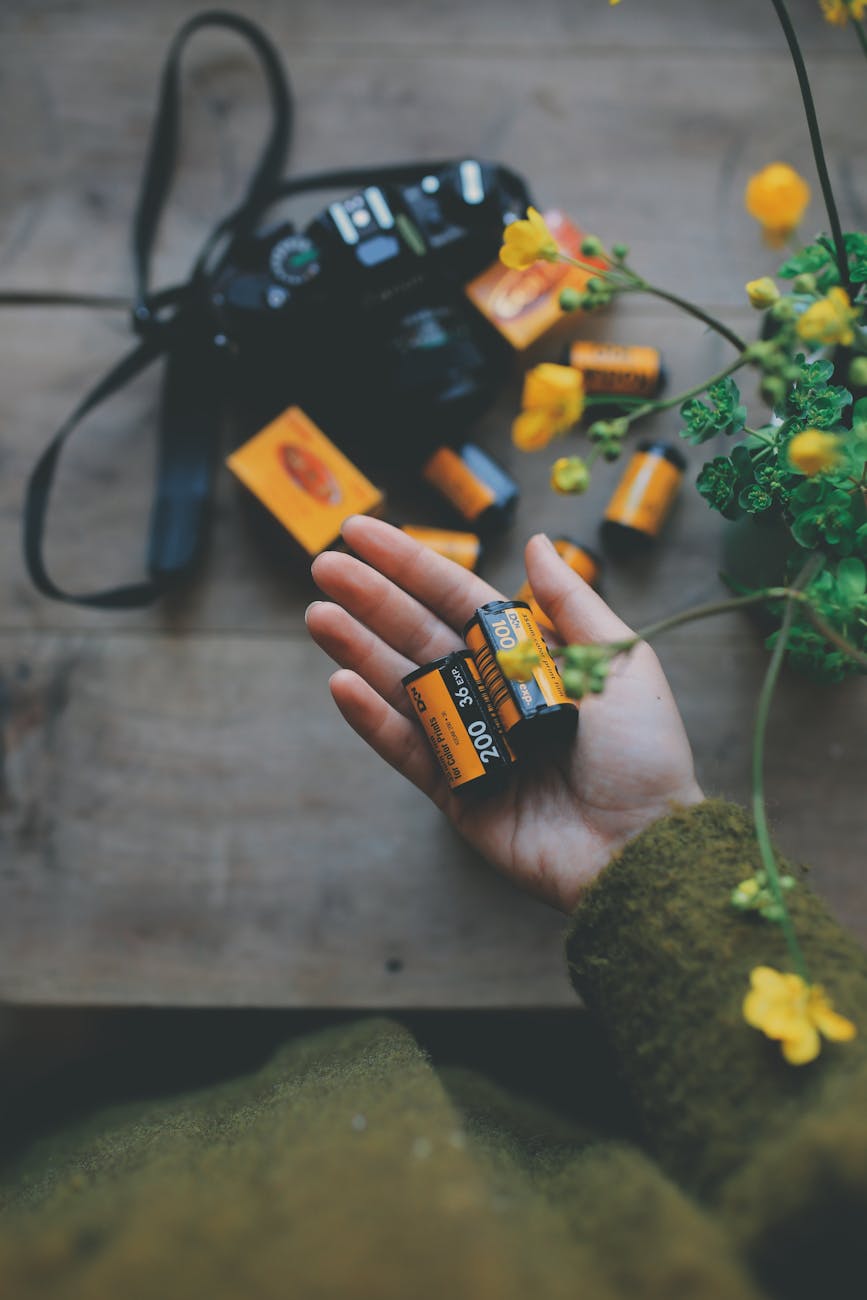 A hand holding film rolls with a vintage camera and yellow flowers beside on a wooden table.