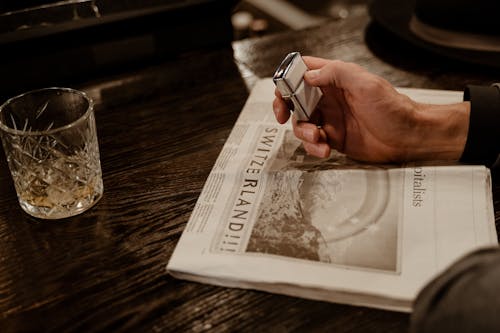 Person Holding Lighter on White Newspaper Beside Empty Glass