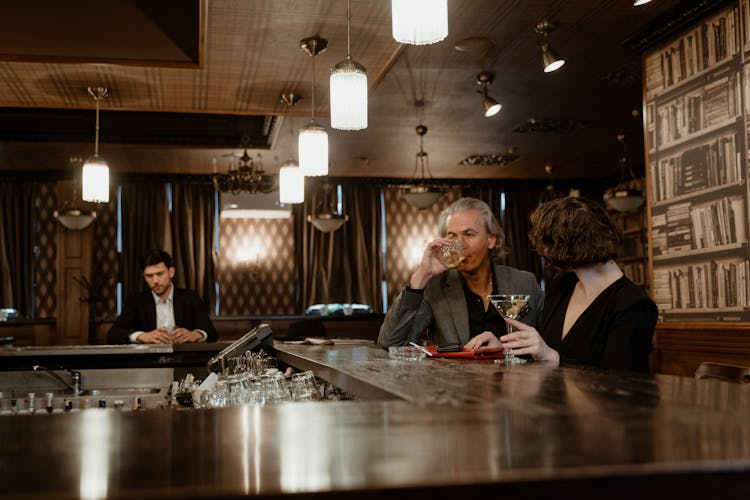 A Mature Man Drinking In A Bar Beside A Person In Black Long Sleeve Shirt