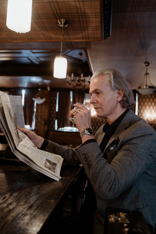 Free Elegant adult man in a gray suit sitting at a bar, reading a newspaper and enjoying a drink in a cozy atmosphere. Stock Photo