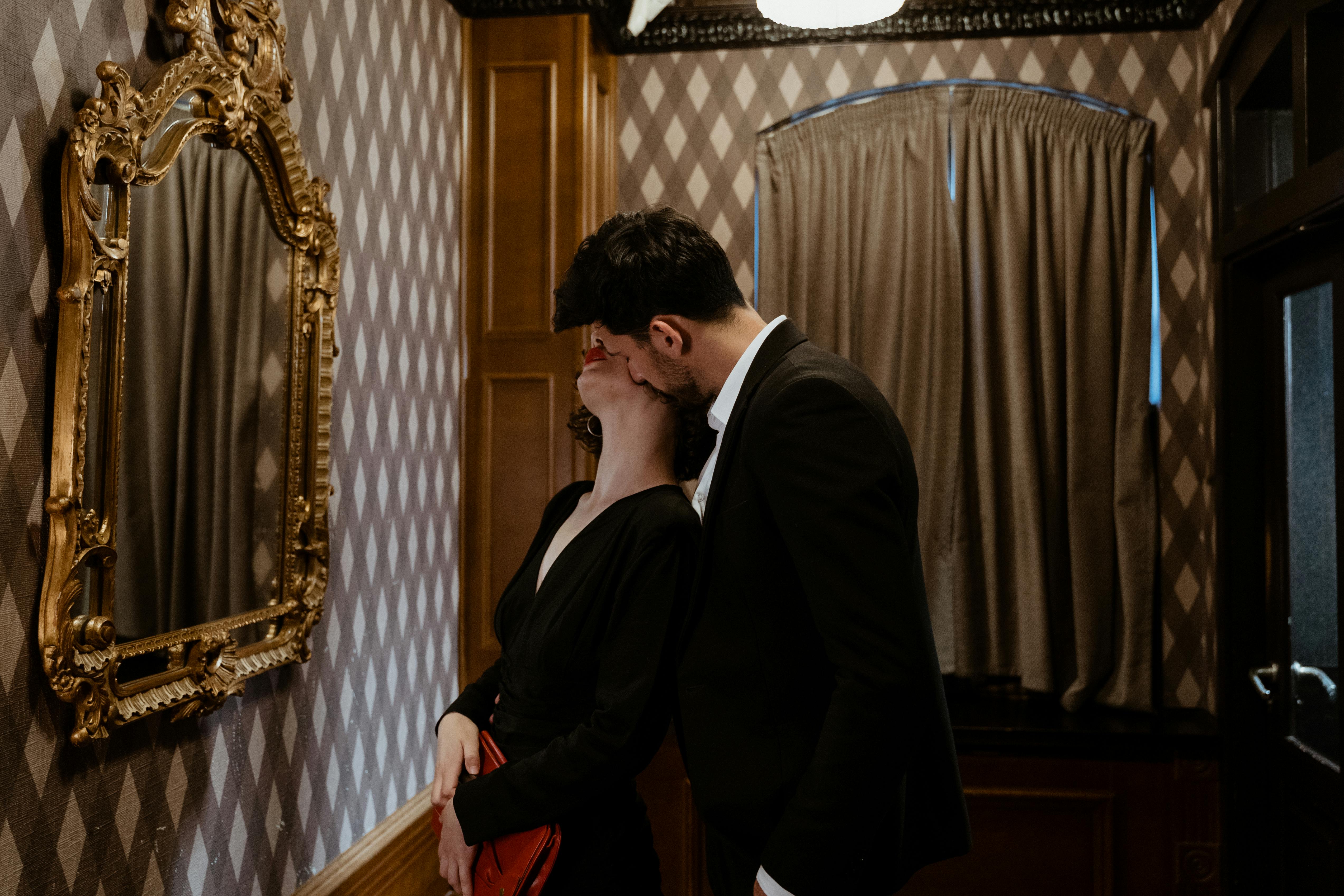 man in black suit kissing woman in black dress kissing on the neck in front of mirror