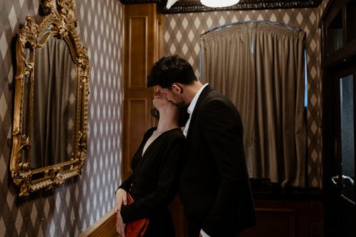 Man in Black Suit Kissing Woman in Black Dress Kissing on the Neck in Front of Mirror