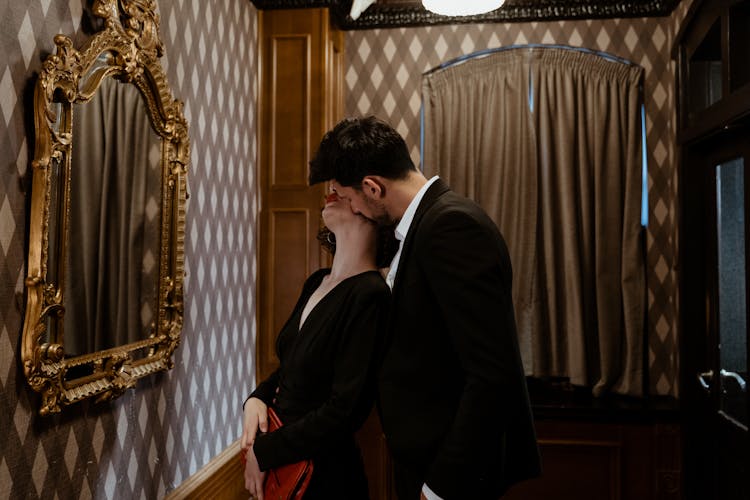 Man In Black Suit Kissing Woman In Black Dress Kissing On The Neck In Front Of Mirror