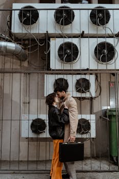 A couple romantically embracing near industrial air conditioners, creating a cinematic mood.