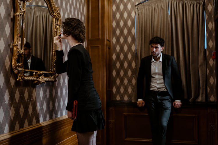 A Man Looking At A Woman Putting Lipstick In Front Of A Mirror