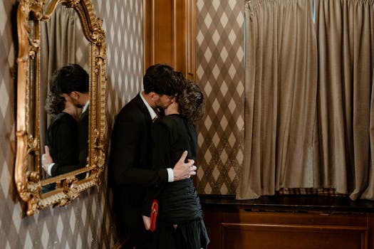 An elegantly dressed couple shares a romantic kiss before an ornate mirror indoors.