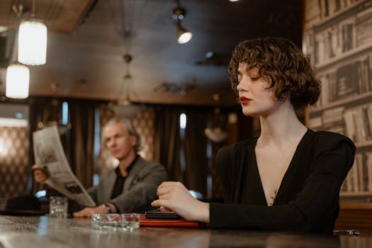 A woman in black attire sitting in a stylish, dimly lit restaurant with a man in the background.