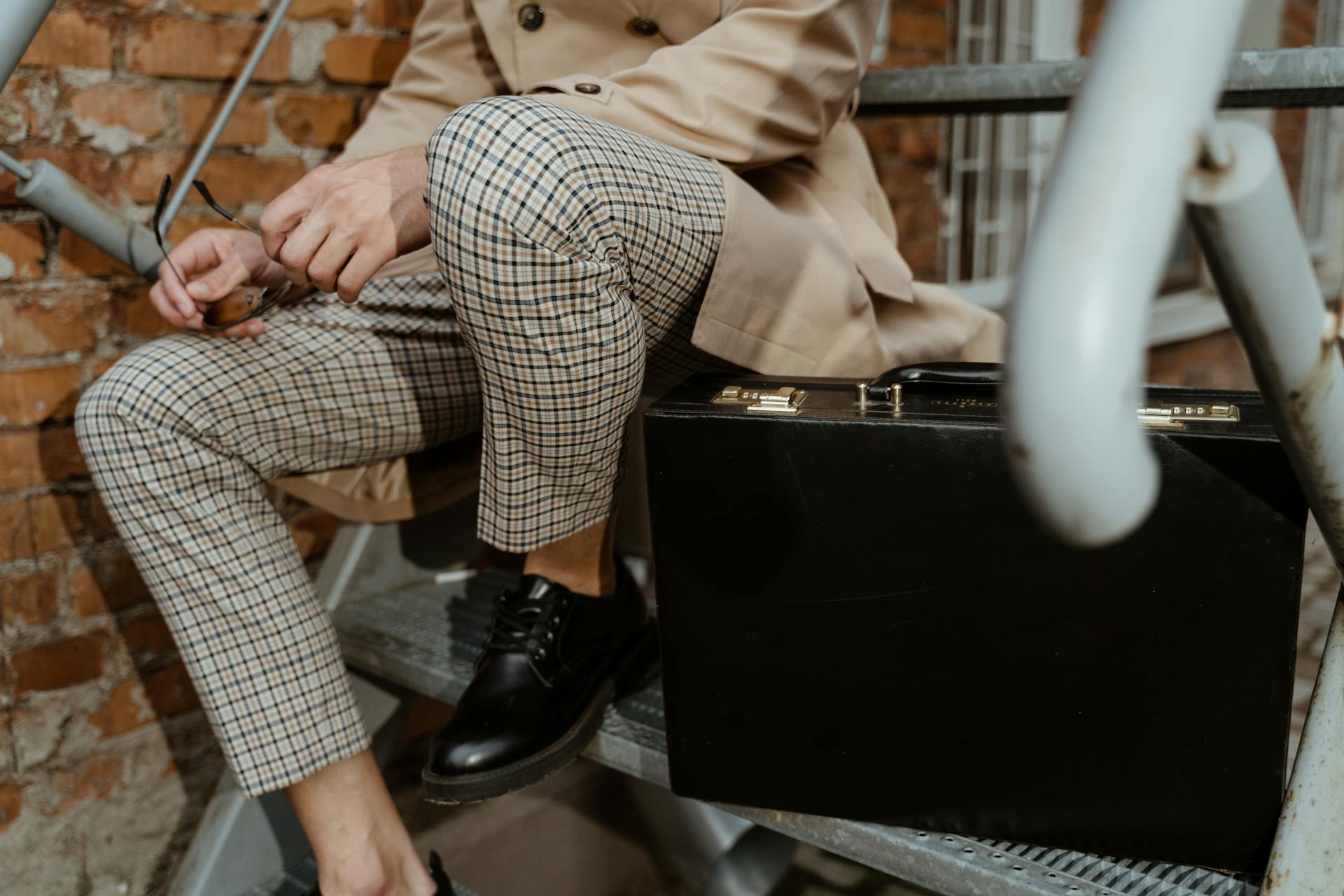 Stylish man wearing plaid pants and holding a briefcase sitting on industrial stairs.