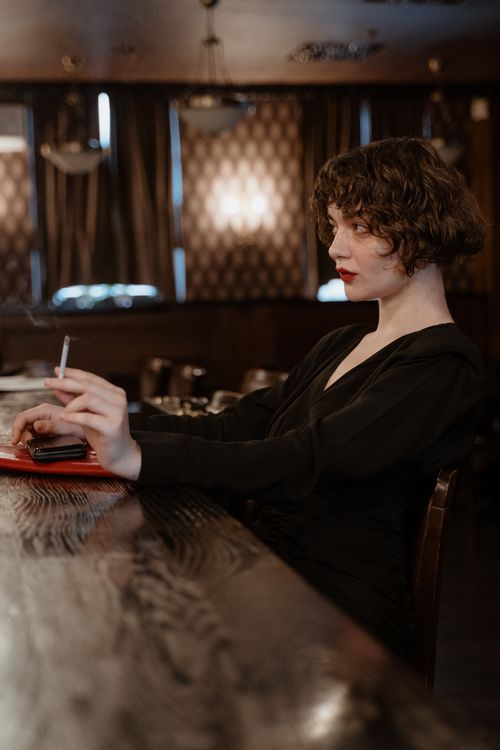 Free Stylish woman sitting indoors holding a cigarette in a dimly lit atmosphere. Stock Photo
