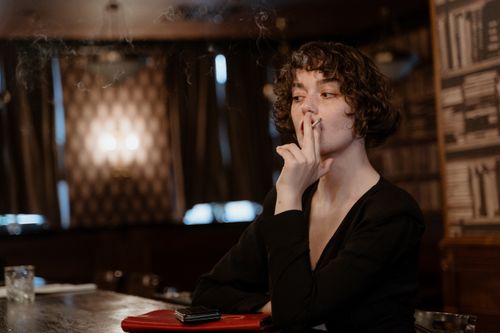 Free A fashionable woman enjoys a cigarette in a cinematic, dimly-lit bar with a classic vibe. Stock Photo