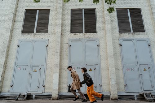 Free A couple in stylish outfits walks past a large industrial brick building. Stock Photo